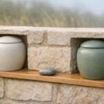 Two matching unmarked ceramic keepsake vessels displayed separately in limestone garden niches, linked by a small river stone and oak detail.